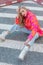 Portrait of striking blonde in bright blouse in coral orange tones, jeans and high shoes, sitting on asphalt in city.
