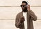 Portrait stylish african man with phone in wireless headphones listening to music wearing brown knitted cardigan and sunglasses