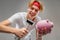 Portrait of a stylish charming blond guy smashing a piggy bank with a hammer on a gray background