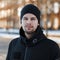 Portrait of a stylish friendly handsome young man with a beard in a fashionable winter black jacket in a black knitted hat on the