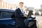 Portrait of a stylish young Caucasian businessman standing near charging electric car, using a laptop computer outdoors