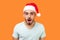 Portrait of surprised bearded young man in santa claus hat and casual white t-shirt standing with open mouth, looking serious and