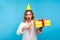 Portrait of surprised beautiful woman with birthday party cone on head pointing at present box and looking with amazement. blue