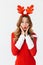 Portrait of surprised woman 20s wearing Santa Claus red costume and deer ears smiling and standing, isolated over white background