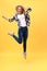 Portrait of surprised young woman in black pants jumping in front of yellow wall. Indoor portrait of young lady in