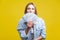 Portrait of surprised young woman in denim shirt peeking out of dollars fan. indoor studio shot  on yellow background