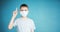 Portrait of a teenager in a gray t-shirt. Photo of a young boy on a blue background. He is in a medical mask, his index finger is