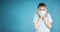 Portrait of a teenager in a gray t-shirt. Photo of a young boy on a blue background. He is in a medical mask, scared, hands