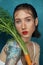 Portrait of tender female fashion model sitting at the blue table with carrot plant. Freckled girl with tattoo directly look at