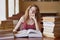 Portrait of thoughtful dreamy young student sitting in reading hall, putting her hand close to face, touching huge opened book,