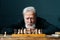 Portrait of thoughtful gray-haired senior older male thinking game strategy sitting on wooden table with chess board