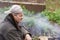 Portrait of thoughtful senior man making bonfire on the backyard. Poor pensioner thinks about his life in smoke screen.