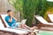 Portrait of tired handsome bearded young adult student man in blue t-shirt and shorts lying on daybed with laptop on poolside and