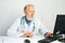 Portrait of tired mature adult male doctor wearing white coat with stethoscope working on desktop computer sitting at