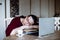 Portrait of tired middle-aged woman sitting at table near laptop, sleeping on crossed arms on pile of book sat home.