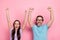 Portrait of two astonished satisfied partners raise fists shout yeah success isolated on pink color background