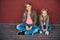 Portrait of two cute little sisters dressed in trendy clothes leaning on a wall while sitting on a skateboard at a