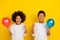 Portrait of two excited positive siblings hold air balloon point finger isolated on yellow color background