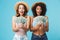 Portrait of two excited women with different type of skin wearing straw hats and summer clothing looking at lots of money holding