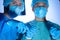 Portrait of two surgeons in sterile gowns, hats and masks. One surgeon, preparing to operate, holds a scalpel with a needle holder