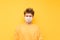 Portrait of upset guy in orange sweatshirt and white medical mask isolated on yellow background, looking down with sad face.