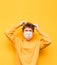 Portrait of an upset shaggy guy in a medical mask on a yellow background, touches his long regrown hair and looks up with a sad