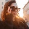 Portrait of an urban stylish woman hipster in trendy sunglasses in a stylish black coat on the background of bright orange sun