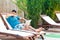 Portrait of wild angry bearded young adult boss man in blue t-shirt and shorts lying on cozy daybed with laptop on poolside and