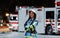 A portrait of a woman in a firefighter uniform with crossed arms, background of a nighttime street where a fire truck is