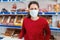 A portrait of woman in a medical mask, with a malicious expression on her face. In the background are shelves of the store. The