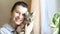 Portrait of a woman with a striped cat at the window of a house with curtains and a ficus houseplant. They look into the frame