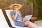 Portrait of wondering serious young adult businesswoman in hat and dress sitting on cozy daybed with laptop and thinking about