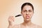 Portrait of a young angry woman applying a skin care mask with a brush. Beige background. Copy space. Concept of cosmetology,