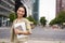 Portrait of young asian woman, looking happy and confident, going to work or university, city skyscrappers behind her