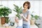 Portrait of young Asian woman smiling friendly holding flower pot with green plant house and looking at camera in living room.