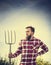 Portrait of young bearded farmer in red checkered shirt with old pitchfork on sky nature backgrund, toned