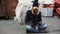 Portrait of young bearded homeless man sitting on a sidewalk near shopping cart ang garbage container during cold winter