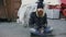 Portrait of young bearded homeless man sitting on a sidewalk near shopping cart ang garbage container during cold winter