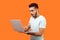 Portrait of young bearded man shocked by message on laptop. indoor isolated on orange background