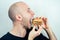 Portrait of a young beautiful bald man eats bites a high-calorie big burger in his hands. concept of diet, unhealthy and