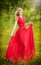 Portrait of young beautiful blonde woman wearing a long red elegant dress posing in a green meadow. Fashionable attractive