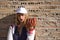 Portrait of young, beautiful, blonde woman with white cap, jacket, baseball glove and ball, leaning against a broken brick wall in