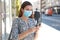 Portrait of young beautiful business woman wearing surgical mask using smart phone app in city street