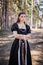 Portrait of a young, beautiful woman in a black medieval dress standing in a pine forest. Attractive girl in a gothic dress.