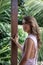 Portrait of young beautiful woman in pink dress with open back on balcony of tropical bungalow with palm trees view