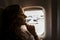 Portrait of young beautiful woman smiling, sitting indoor and looking forward on background of airplane window with airport view.