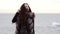 Portrait of a young brunette woman in fur coat standing by the sea in winter while the wave hits the ground and causes