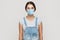 Portrait of young brunette woman with surgical medical mask in denim overalls standing and looking at camera. medical and health