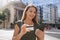 Portrait of young business woman watching her phone holding credit card in finance district avenue