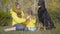 Portrait of young Caucasian woman sitting on green grass and putting mustard hat on girl. Beautiful graceful doberman